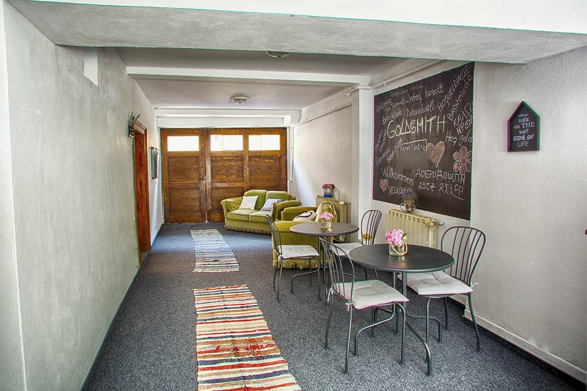 Gangul boltit dintre poartă și curte, cu preșuri țesute pe jos, canapele verde-măsliniu, mese bistro și tabla mare de scris pe care este scris cu cretă GOLDSMITH, înconjurat de urări de bun venit în mai multe limbi.